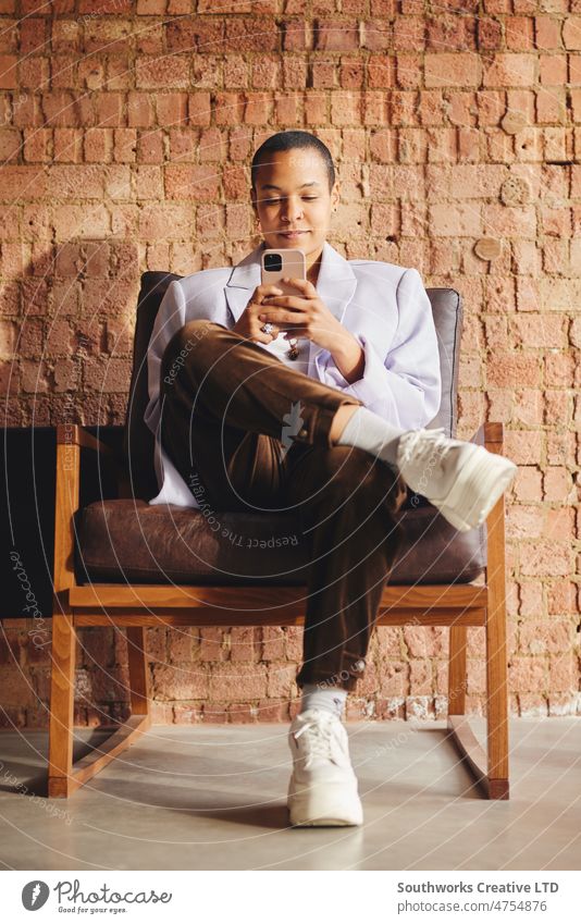 Porträt einer selbstbewussten multirassischen LGBTQ-Frau im mittleren Erwachsenenalter, die auf einem Retro-Stuhl sitzt und ein Smartphone vor einer freiliegenden Backsteinwand benutzt