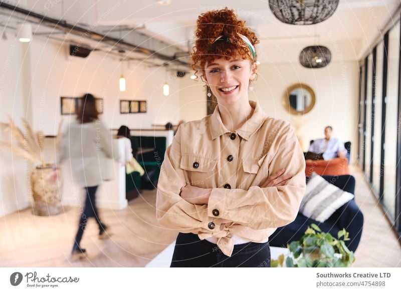 Porträt einer selbstbewussten jungen weißen Frau mit roten lockigen Haaren, die lächelnd und mit verschränkten Armen in die Kamera schaut, in einem Coworking Space