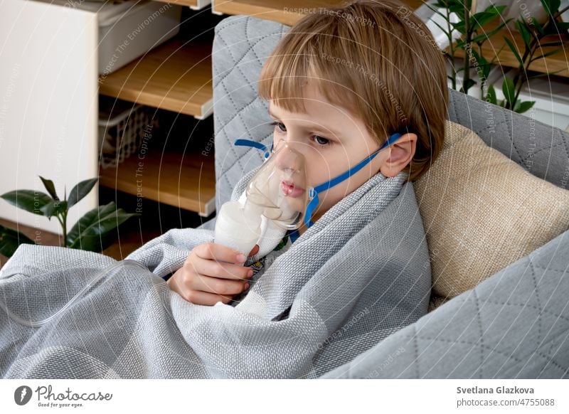 Kleiner kaukasischer Junge, der zu Hause mit einem Vernebler inhaliert. Kind hält einen Maskendampf-Inhalator in der Hand. Behandlung von Asthma. Konzept eines Inhalationstherapiegerätes.