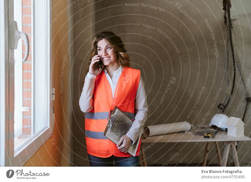 Professionelle Architektin auf einer Baustelle, die mit einem Mobiltelefon spricht und Baupläne hält Frau Blaupausen Arbeitsbereich Schutzhelm Schutzmantel