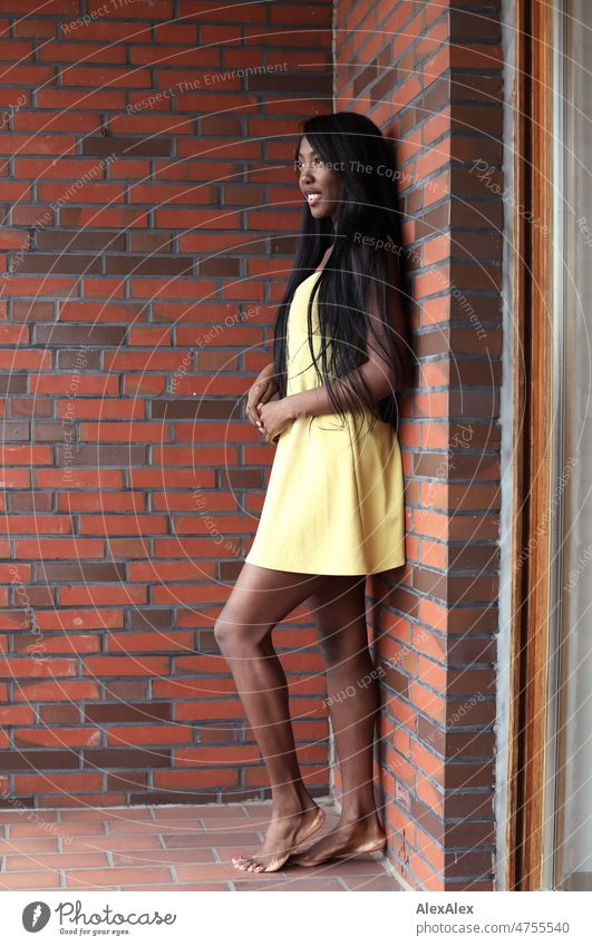 Junge, schöne, langhaarige Frau in gelbem Sommerkleid steht barfüßig auf dem Balkon vor Ziegelsteinwand und lächelt junge Frau Schönheit Anmut Freude