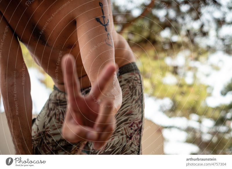 Junger Mann in Badehose zeigt Friedenszeichen mit Fingern im Schatten eines Baumes jung mann friede Friedenssymbole friedenszeichen Männer Männlichkeit Herz