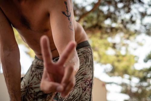 Junger Mann in Badehose zeigt Friedenszeichen mit Fingern im Schatten eines Baumes jung mann friede Friedenssymbole friedenszeichen Männer Männlichkeit Herz