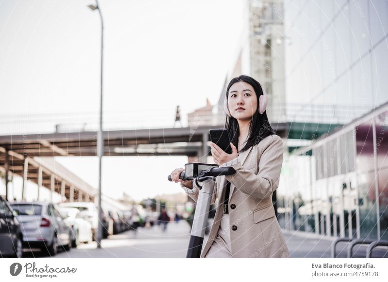 Chinesische Geschäftsfrau, die einen Elektroroller in der Stadt mietet, Kopfhörer trägt und ein Mobiltelefon benutzt Frau elektrisch Tretroller Musik Großstadt