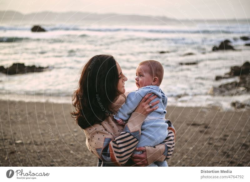 Alleinstehende junge Mutter hält ihr Baby am Strand und schaut es an Stille sanft heiter Tante Sweatshirt Meeres-Hintergrund Eltern Himmel Frau Kind Familie