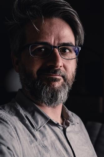Portrait von einem Mann mit Brille und Bart, der zaghaft lächelt dunkel Hemd schmunzeln lächeln Blick in die Kamera
