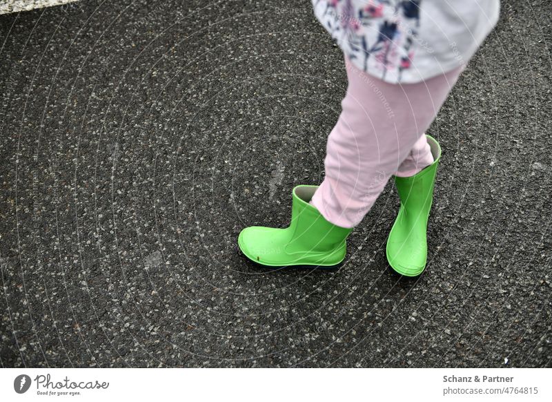 Kind mit grünen Gummistiefeln und verschränkten Beinen Stiefel Wetter Asphalt Schuhe über Kreuz Hose rosa spielen Straße stehen