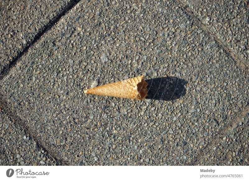 Leere angeknabberte Waffel einer Eistüte mit Licht und Schatten bei Sonnenschein auf grauem Straßenpflaster im Frühling in der Fußgängerzone in Detmold in Ostwestfalen-Lippe