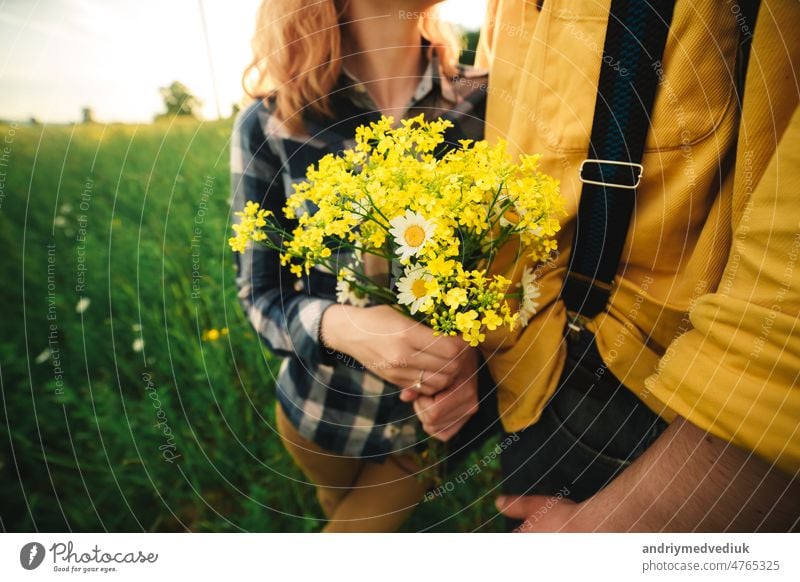 ausgeschnittenes Foto von Blumenstrauß von Wildblumen in der Hand der Frau. Verliebtes Hipster-Paar, das im Feld spazieren geht, sich küsst und die Hände hält, sich umarmt und im Sommer bei Sonnenuntergang im Gras liegt. valentines day