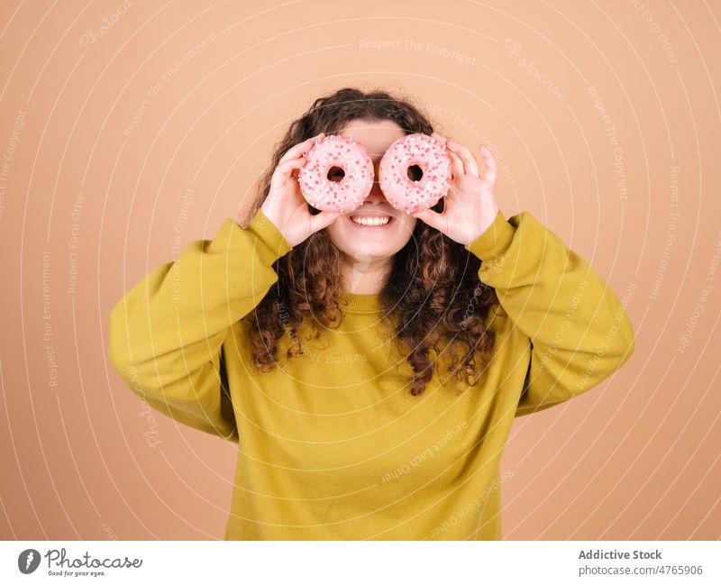 Fröhliche Frau, die sich mit süßen Donuts im Studio die Augen zuhält Model Glasur Spaß haben Glück Vergnügen bestäuben Augen abdecken heiter positiv Dessert