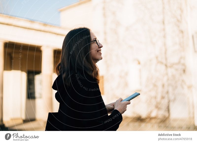 Lächelnde Frau mit Brille und schwarzem Kapuzenpulli, die im Freien ihr Smartphone benutzt positiv benutzend Anschluss Nachricht tausendjährig Browsen Großstadt