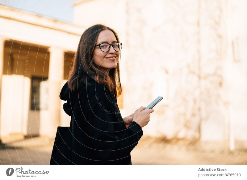 Lächelnde Frau mit Brille und schwarzem Kapuzenpulli, die im Freien ihr Smartphone benutzt positiv benutzend Anschluss Nachricht tausendjährig Browsen Großstadt