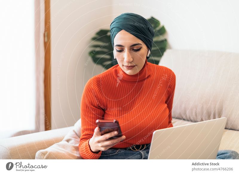 Muslimische Frau benutzt Smartphone auf Sofa Porträt Laptop Kopfhörer benutzend Ohrstöpsel tws Drahtlos heimwärts ruhen zuhören Musik soziale Netzwerke