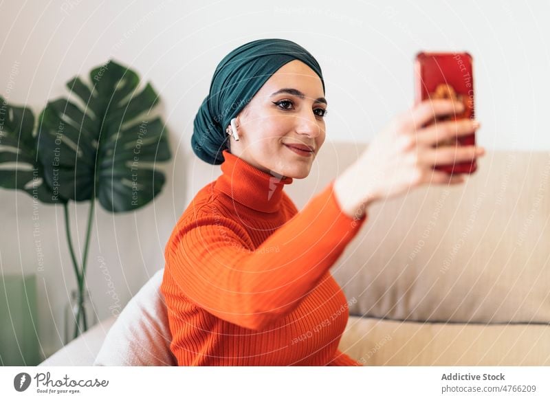 Muslimische Frau macht Selfie zu Hause Porträt heimwärts Wohnzimmer Smartphone Lächeln Sofa Musik zuhören Wochenende muslimisch ethnisch Islam Kopftuch
