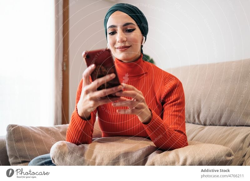 Muslimische Frau benutzt Smartphone auf Sofa Porträt benutzend heimwärts ruhen Kopfhörer Ohrstöpsel tws Drahtlos zuhören Musik soziale Netzwerke Wochenende