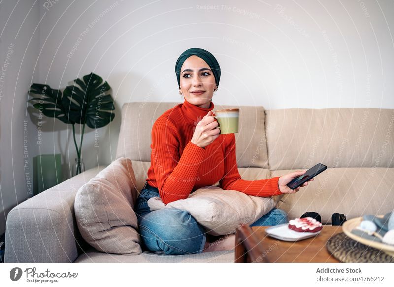 Muslimische Frau genießt ein heißes Getränk auf dem Sofa Porträt Heißgetränk ruhen Wochenende heimwärts Smartphone Lächeln Wohnzimmer froh muslimisch ethnisch