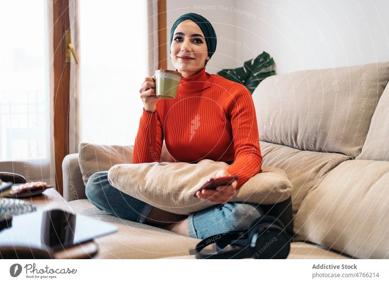 Muslimische Frau genießt ein heißes Getränk auf dem Sofa Porträt Heißgetränk ruhen Wochenende heimwärts Smartphone Lächeln Wohnzimmer froh muslimisch ethnisch