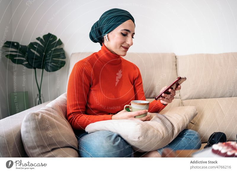 Muslimische Frau genießt ein heißes Getränk auf dem Sofa Porträt Heißgetränk ruhen Wochenende heimwärts Browsen Smartphone Kopfhörer Ohrstöpsel Lächeln Drahtlos