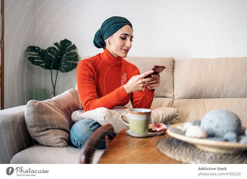 Muslimische Frau genießt ein heißes Getränk auf dem Sofa Porträt Heißgetränk ruhen Wochenende heimwärts Browsen Smartphone Kopfhörer Ohrstöpsel Lächeln Drahtlos