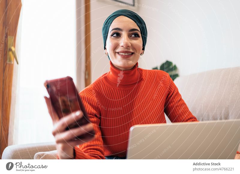 Muslimische Frau benutzt Smartphone auf Sofa Porträt Laptop heimwärts ruhen soziale Netzwerke muslimisch ethnisch Islam lässig Kopftuch Wohnzimmer Apparatur
