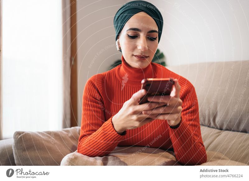 Muslimische Frau benutzt Smartphone auf Sofa Porträt benutzend heimwärts ruhen Kopfhörer Ohrstöpsel tws Drahtlos zuhören Musik soziale Netzwerke Wochenende