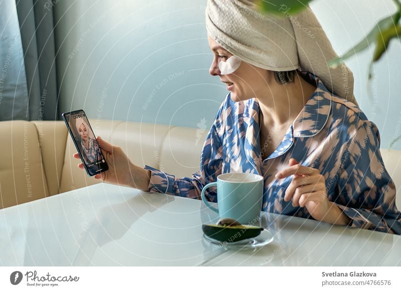 Morgenroutine. Eine Frau in Hauskleidung, Schlafanzug und Handtuch trinkt beim Frühstück einen Kaffee und spricht mit einem Mobiltelefon Smartphone