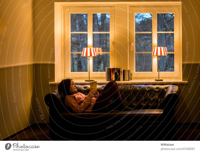 Eine junge Frau liegt gemütlich auf einem Sofa am Fenster und liest, draußen ist blaue Stunde Lesen Bücher Mädchen Couch warm Abend Lampen Tischlampen Licht