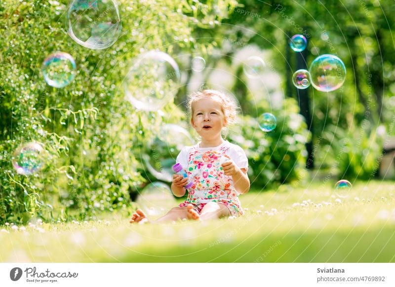 Ein kleines Mädchen pustet im Park Seifenblasen, während es im Gras sitzt. Das Konzept einer glücklichen Kindheit wenig Schaumblase Sommer Glück Blasen niedlich