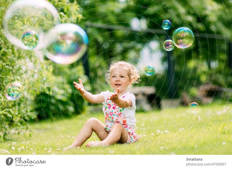 Ein kleines Mädchen fängt Seifenblasen im Park, während es im Gras sitzt. Das Konzept einer glücklichen Kindheit wenig Schaumblase Sommer Glück Blasen niedlich