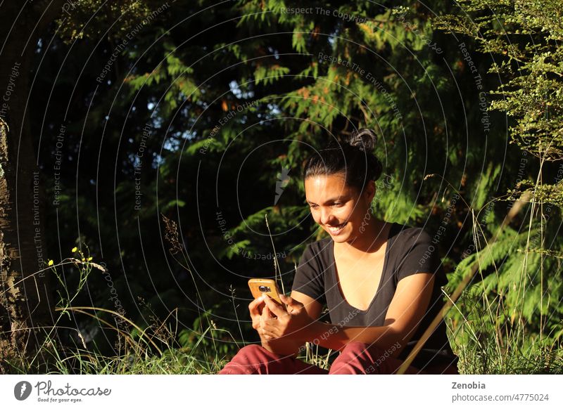 Frau sitzt draußen in der Natur, schaut auf das Telefon und lächelt. Erwachsener attraktiv schön schwarz braun Brötchen Kaukasier Mitteilung Anschluss dunkel
