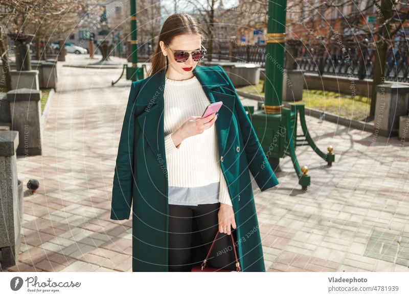 Junge brünette Frau in legerem Mantel schreibt online SMS mit Smartphone beim Chatten auf der Straße urban Telefon Mode jung Großstadt Mädchen Mobile