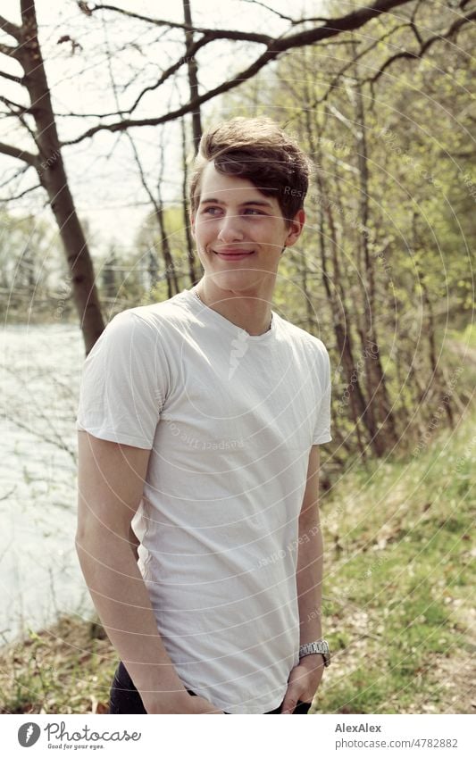 Junger Mann steht an einem Seeufer und schaut lächelnd zur Seite junger Mann Jugendlicher draussen Natur groß schön sportlich dunkelblond T-Shirt weißes Shirt