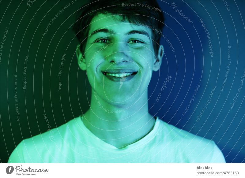 Portrait eines jungen Mannes mit Wangengrübchen in grünem und blauem Licht vor petrol-farbener Wand Junge junger Mann Jugendlicher groß schön stark sportlich