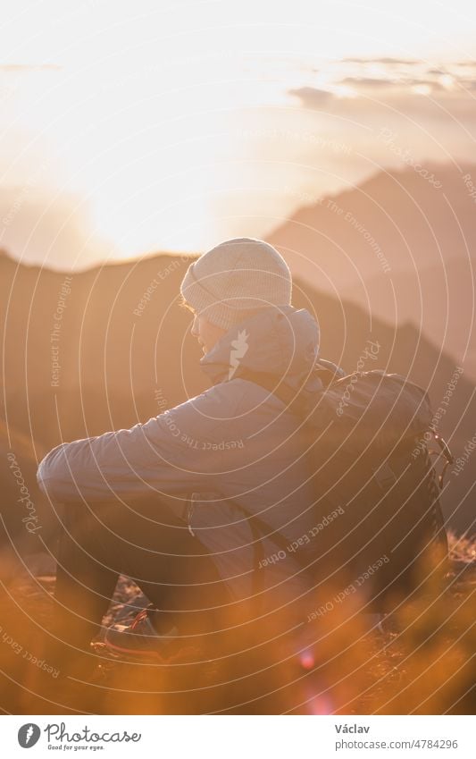 Junger Abenteurer, der bei Sonnenaufgang am Rande eines Felsens auf dem höchsten Berg Pico Ruivo auf der Insel Madeira, Portugal, sitzt. Das orange-goldene Licht beleuchtet den jungen Mann. Unverfälschtes Porträt