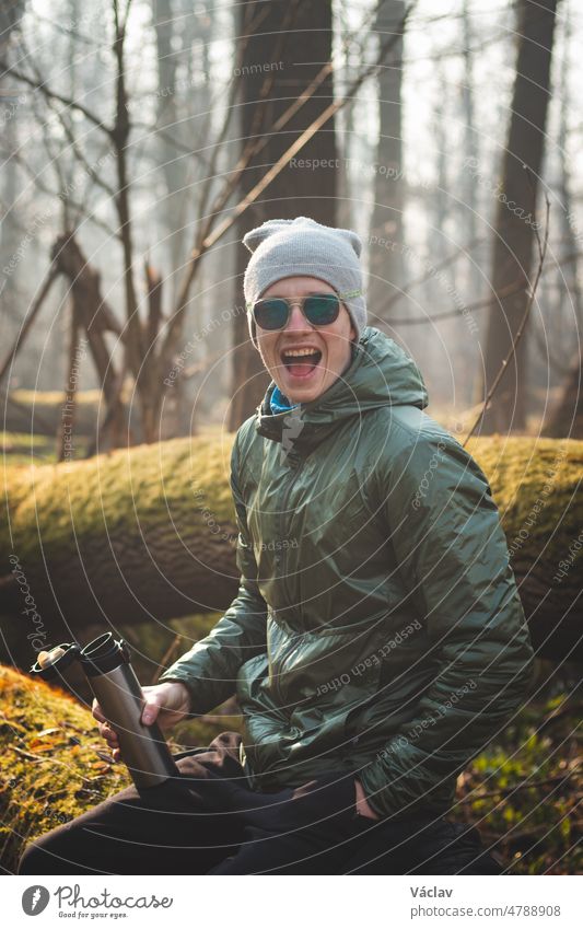 Lachender, erfahrener Wanderer, der in der Wildnis auf einem Baumstamm sitzt und seinen heißen Tee aus einer Thermoskanne trinkt. Echte Menschen, die die Artenvielfalt der tschechischen Wälder entdecken. Ein echtes Lächeln