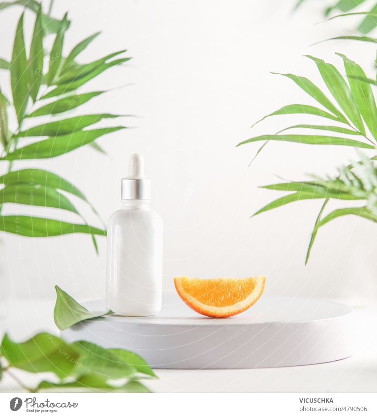 Kosmetische Pipettenflasche und Orangenscheibe auf Podest vor weißem Vitrinenhintergrund mit grünen tropischen Blättern. Kosmetik orange Scheibe Podium