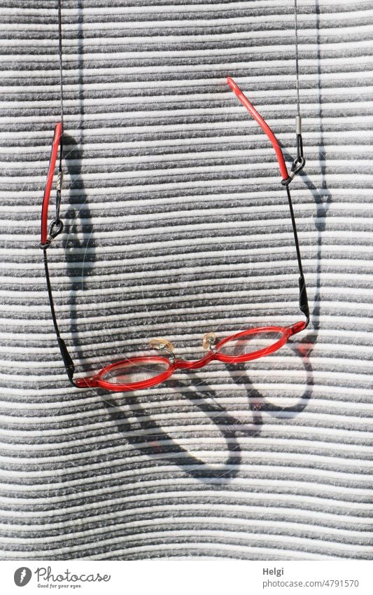 Brillenorden am Bande - rote Brille mit großem Schatten hängt an einem Brillenband vor grau-weiß gestreiftem Pullover | UT Frühlingslandluft Sehhilfe Licht