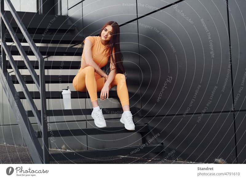 Sitzt auf der Treppe und hält eine Flasche Wasser. Junge Frau in Sportkleidung hat Fitness-Sitzung im Freien jung Erwachsener Körper gelb Kleidung Europäer