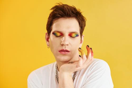 Junger schwuler Mann steht im Studio und posiert für eine Kamera. Regenbogen Make-up jung Porträt Gesicht Geschlecht lgbt Typ Transgender androgyn gelb
