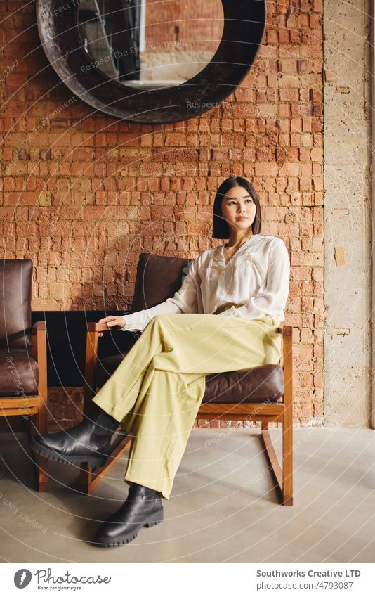 Porträt einer selbstbewussten chinesischen Geschäftsfrau im mittleren Erwachsenenalter, die auf einem Retro-Stuhl in einem Büro mit freiliegender Backsteinwand sitzt