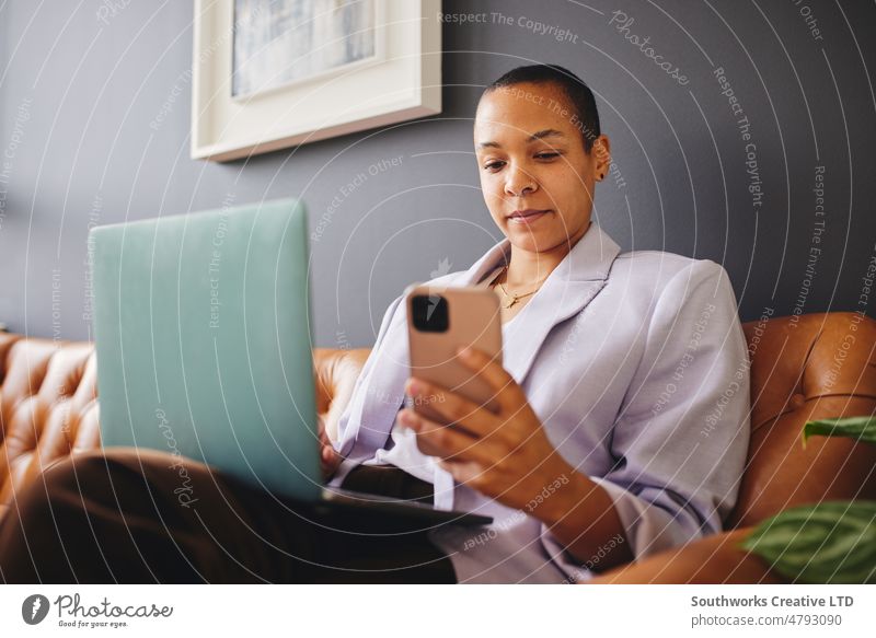 Porträt einer multirassischen LGBTQ-Frau im mittleren Erwachsenenalter, die Smartphone und Laptop auf der Couch benutzt und sich konzentriert rassenübergreifend