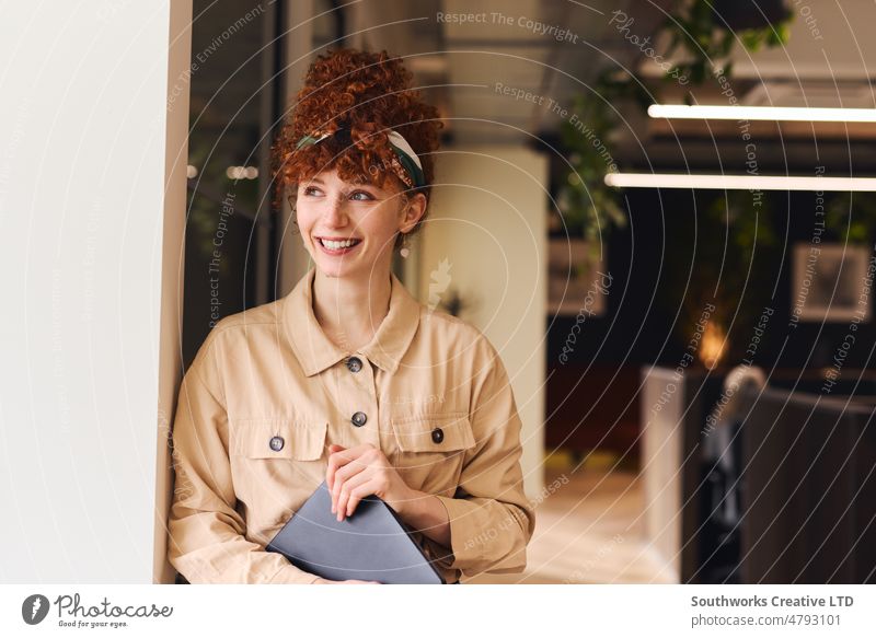 Porträt einer selbstbewussten jungen weißen Frau mit roten lockigen Haaren, die lächelt und wegschaut, während sie einen Laptop in einem Coworking Space hält