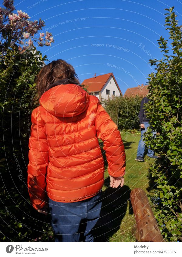 Junge mit langen Haaren und Daunenjacke in leuchtendem Orange bei blauem Himmel und Sonnenschein im Frühling in einem Garten in Wettenberg Krofdorf-Gleiberg bei Gießen in Hessen