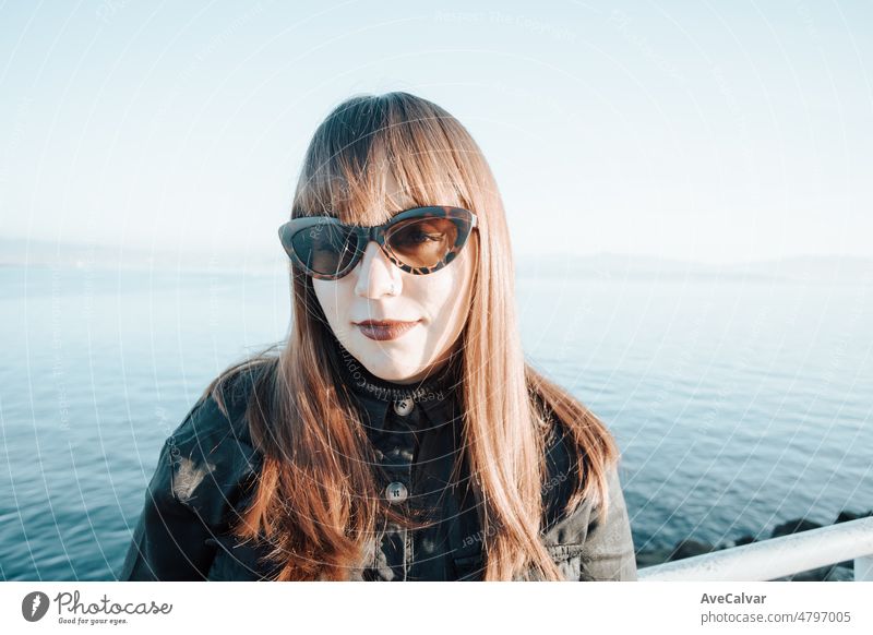 Moderne junge Frau mit Sonnenbrille an einem sonnigen Sommertag mit halbem Lächeln und Blick in die Kamera. Alle schwarzen tragen an heißen Tagen. Urlaub am Mittelmeer für nordische Menschen Konzept.