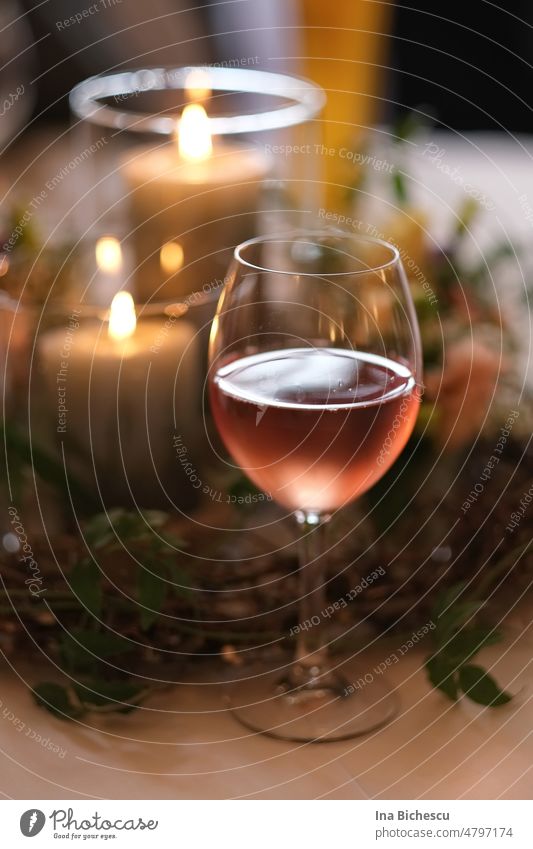 Ein beschlagenes Weinglas halbvoll mit Rosé Wein vor zwei angezündete Kerzen auf unscharfen pflanzliche Dekoration. Glas Beschlagen kalt Roséwein