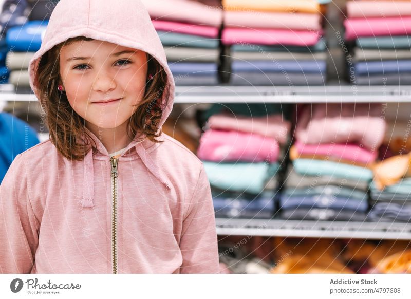 Kleines Mädchen im Kleiderladen Werkstatt Regal Lächeln Kapuzenpulli lässig Kunde froh Porträt niedlich Kind positiv Einzelhandel freundlich Stoff wenig