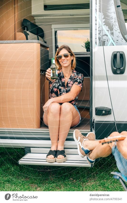 Frau, die eine Bierflasche zeigt, hat Spaß mit Freunden, die an der Tür ihres Wohnmobils sitzen Lächeln zeigend Flasche Kleintransporter Sitzen Glück sprechend