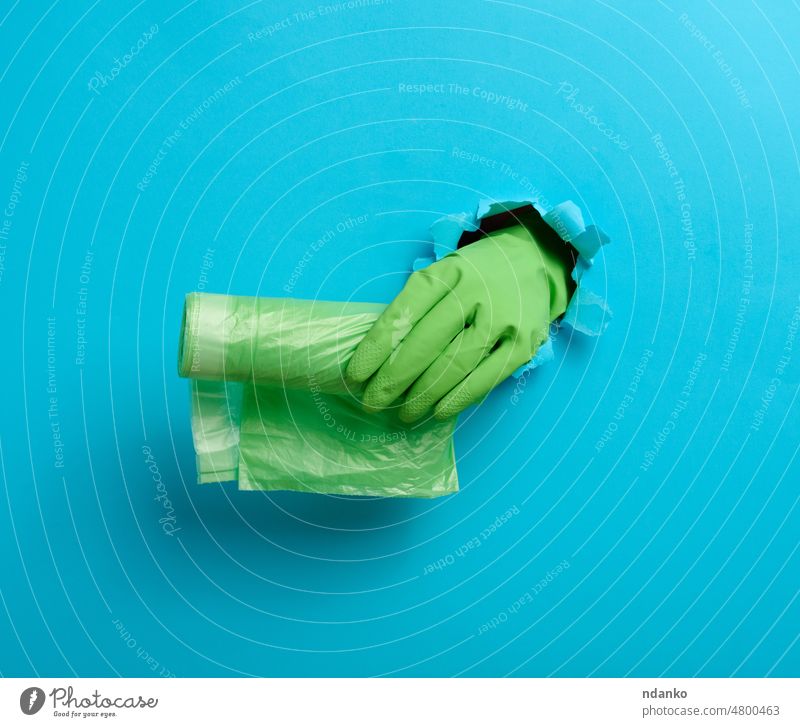 Hand hält ein Bündel transparenter Müllsäcke auf blauem Hintergrund. Ein Teil des Körpers ragt aus einem zerrissenen Loch im Papier heraus Handschuh grün Arme