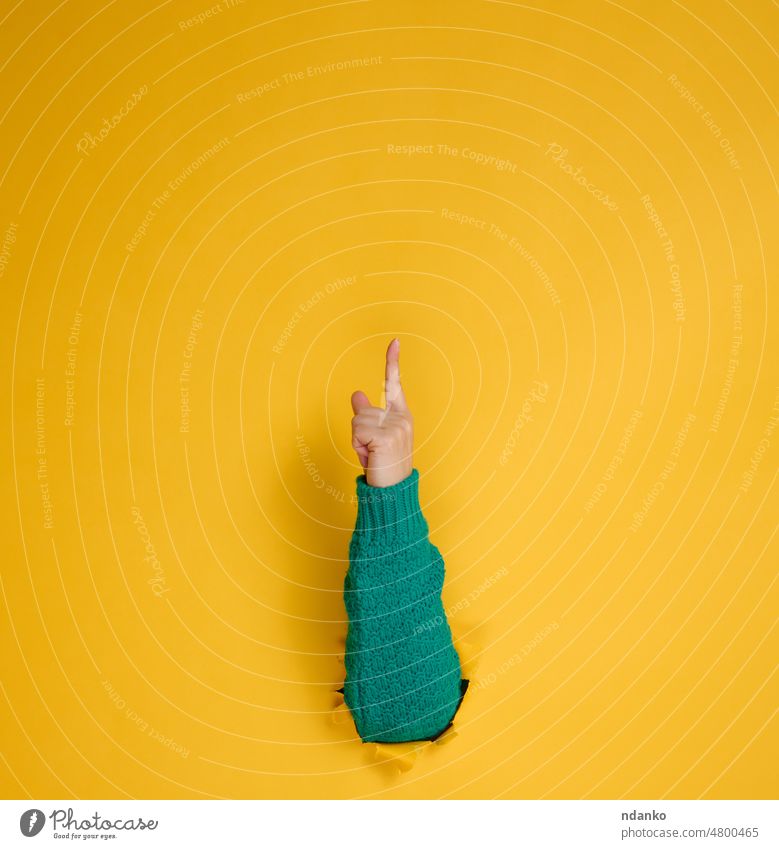 weibliche Hand ragt aus einem zerrissenen Loch in einem gelben Papierhintergrund, ein Teil des Körpers zeigt mit dem Zeigefinger nach oben. Leerzeichen kopieren