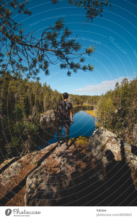 Ein 24-jähriger Reisender liegt auf einem hohen Felsen und beobachtet einen wunderschönen natürlichen See im Hiidenportti-Nationalpark, Sotkamo in der Region Kainuu, Finnland. Wandern in der finnischen Landschaft